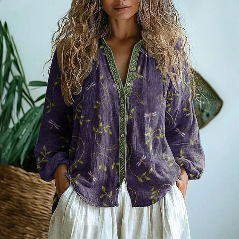 Frau in lila Bluse mit floralem Muster und weißem Rock, vor Pflanzen stehend. Modetrend, Boho-Stil, lässige Damenmode, Sommeroutfit.