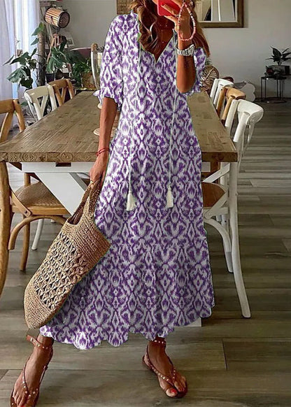 Frau in lila-weißem Boho-Kleid mit Ikat-Muster, hält eine geflochtene Tasche, steht in modernem Esszimmer mit Holztisch und weißen Stühlen.