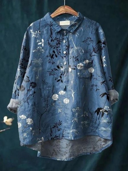 Blaues Hemd mit floralen Mustern, hängend auf einem Holzkleiderbügel vor dunklem Hintergrund. Elegantes Design, modische Damenbekleidung.