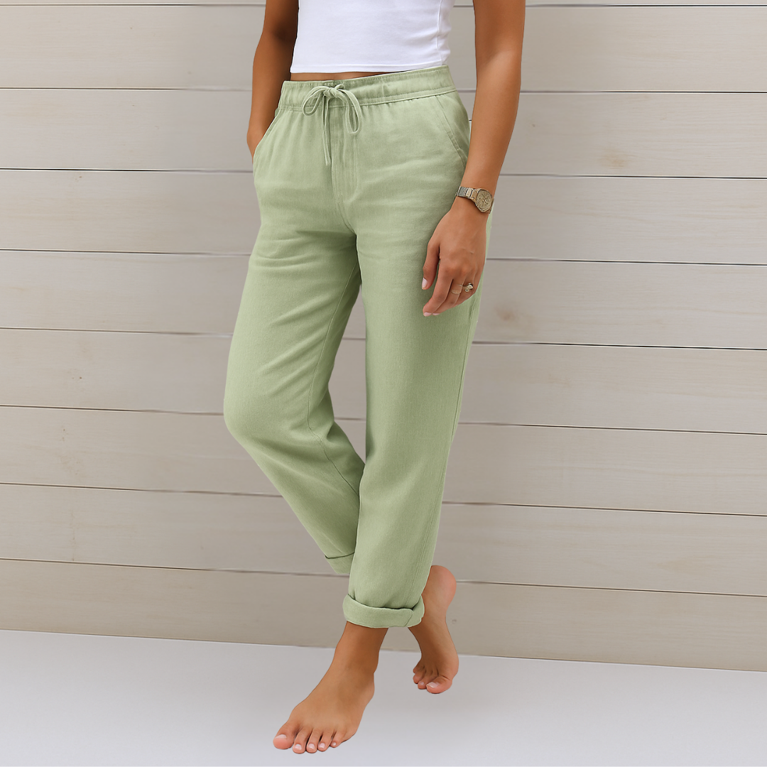Frau in grüner Leinenhose und weißem Top, barfuß vor Holzwand. Modetrend, Sommeroutfit, lässiger Stil, Damenbekleidung.