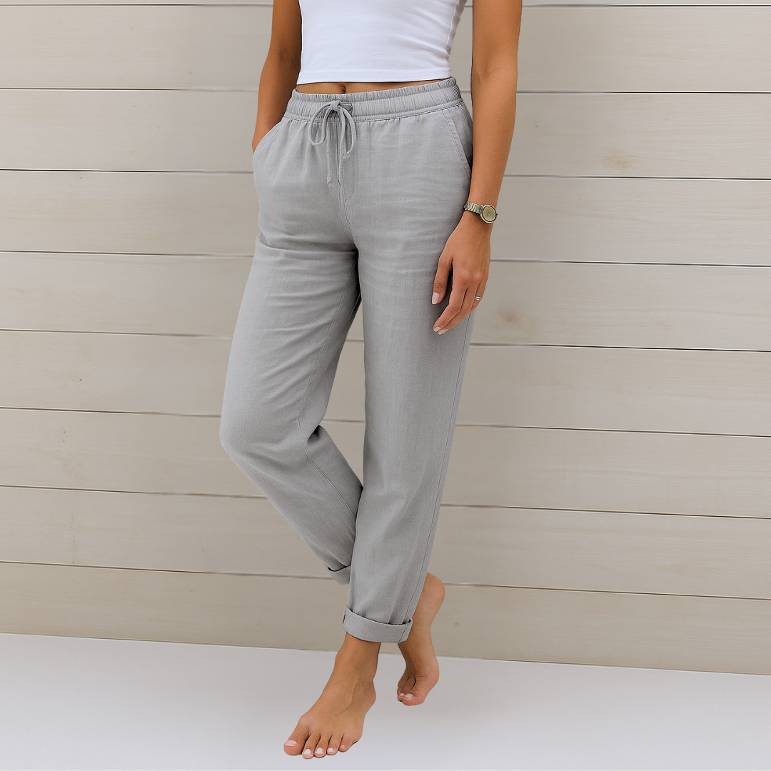 Frau in grauer Leinenhose und weißem Top vor Holzwand. Bequeme Damenmode, lässiger Stil, Sommeroutfit, Freizeitkleidung.