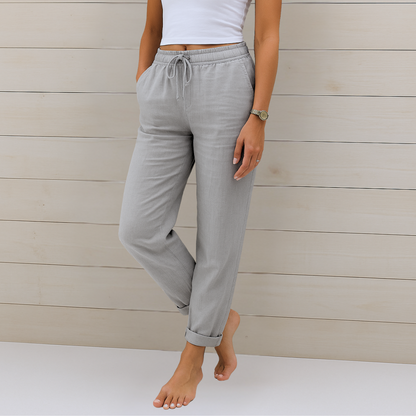Frau in grauer Leinenhose und weißem Top vor Holzwand. Bequeme Damenmode, lässiger Stil, Sommeroutfit, Freizeitkleidung.