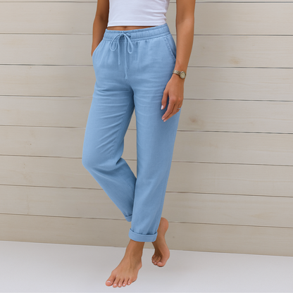 Frau in hellblauer Leinenhose und weißem Top vor Holzwand. Sommermode, bequeme Damenbekleidung, Freizeitlook, stilvolle Leinenhose.