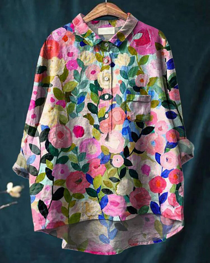 Bunte Damenbluse mit floralem Muster, langen Ärmeln und Knopfleiste, hängend auf einem Kleiderbügel vor dunklem Hintergrund.
