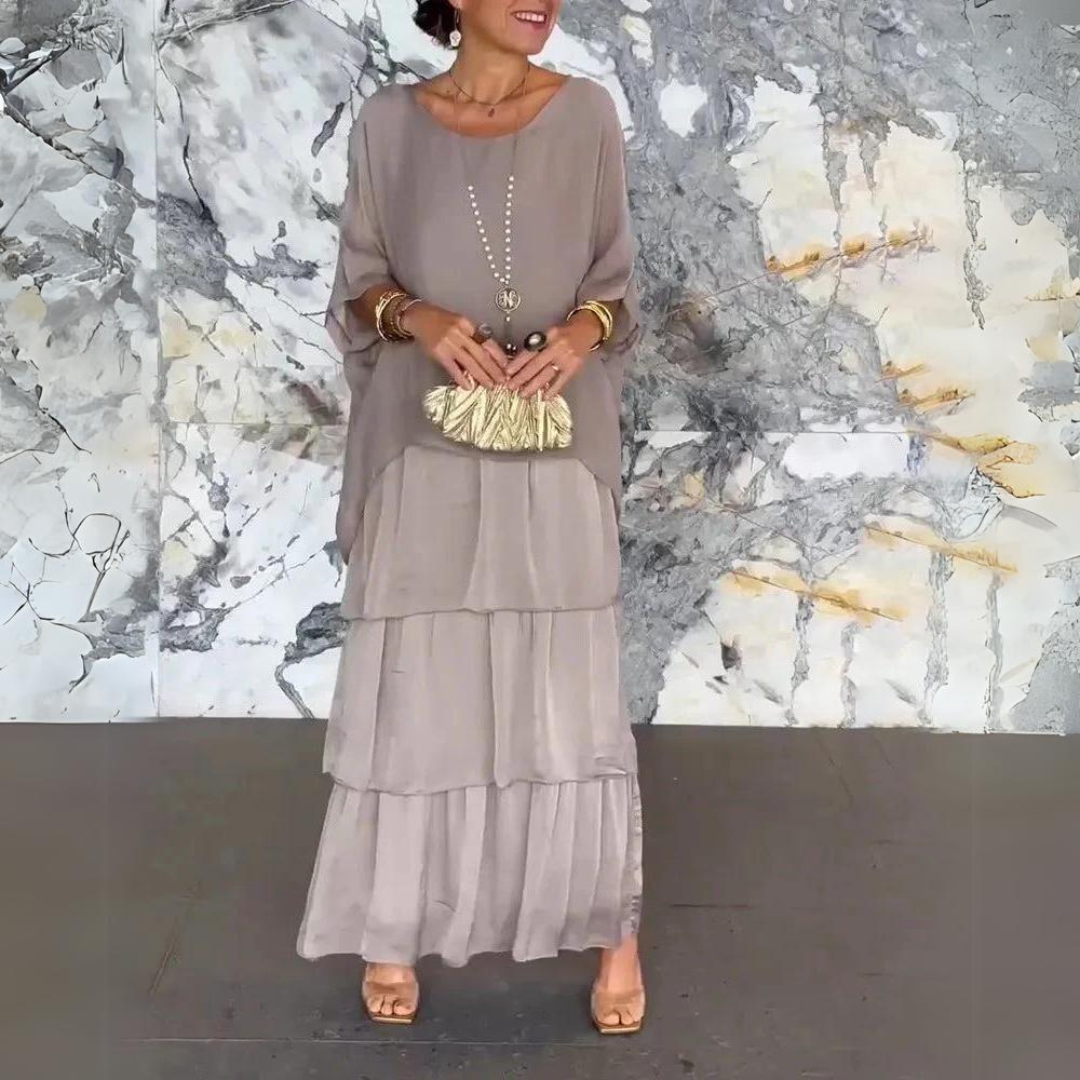 Elegantes Outfit mit grauem, mehrlagigem Kleid und goldener Clutch vor marmorierter Wand. Perfekt für stilvolle Anlässe und Mode-Events.