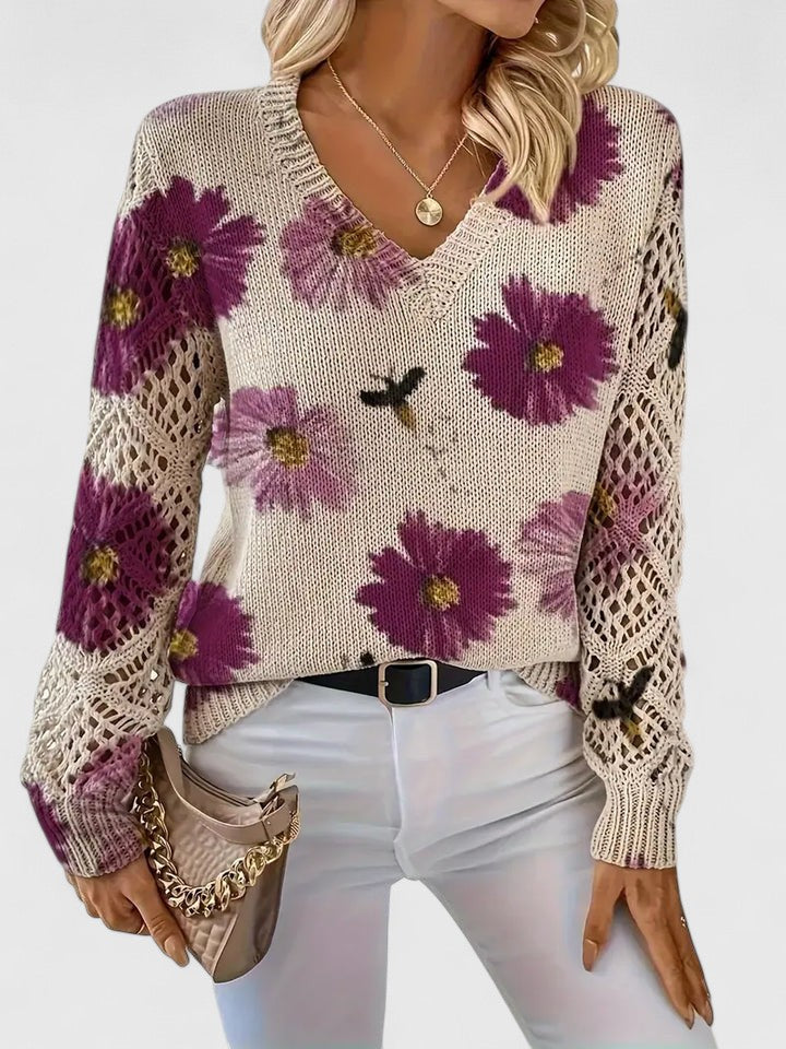 Frau trägt cremefarbenen Pullover mit lila Blumenmuster, V-Ausschnitt und gehäkelten Ärmeln, kombiniert mit weißer Hose und beiger Handtasche.
