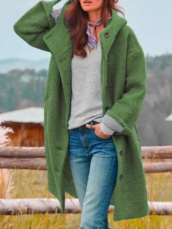 Frau in grünem Wollmantel, grauem Pullover und blauer Jeans steht vor ländlicher Kulisse. Herbstmode, Damenbekleidung, Outdoor-Stil.