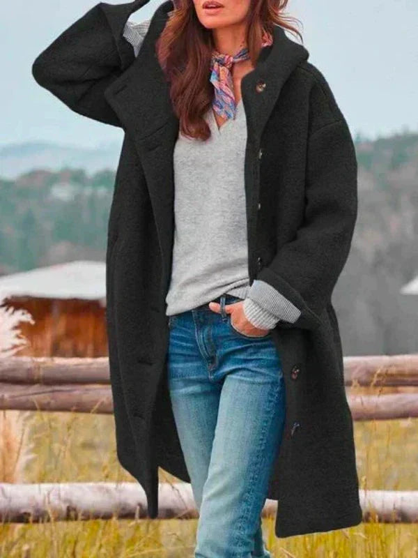 Frau in schwarzem Wollmantel, grauem Pullover und blauer Jeans steht vor ländlicher Kulisse. Herbstmode, Damenbekleidung, Outdoor-Stil.