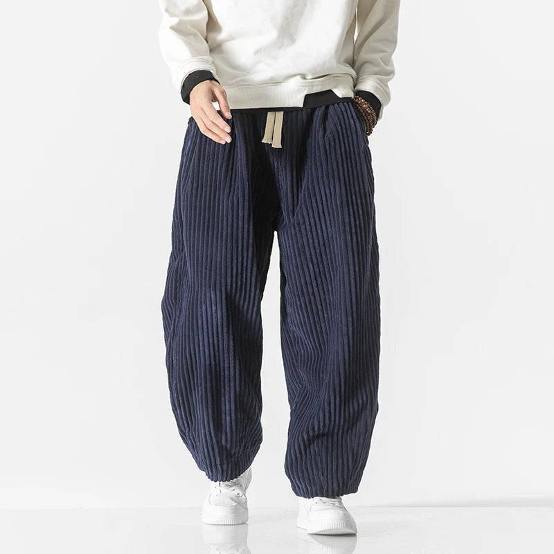 Mann trägt weite, gestreifte Cordhose in Dunkelblau mit weißem Pullover und weißen Sneakers. Modetrend, Herrenbekleidung, lässiger Stil.