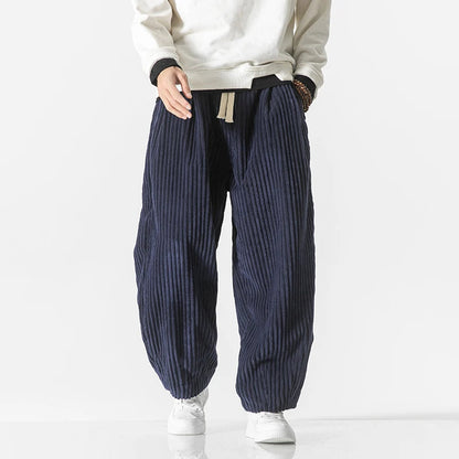Mann trägt weite, gestreifte Cordhose in Dunkelblau mit weißem Pullover und weißen Sneakers. Modetrend, Herrenbekleidung, lässiger Stil.