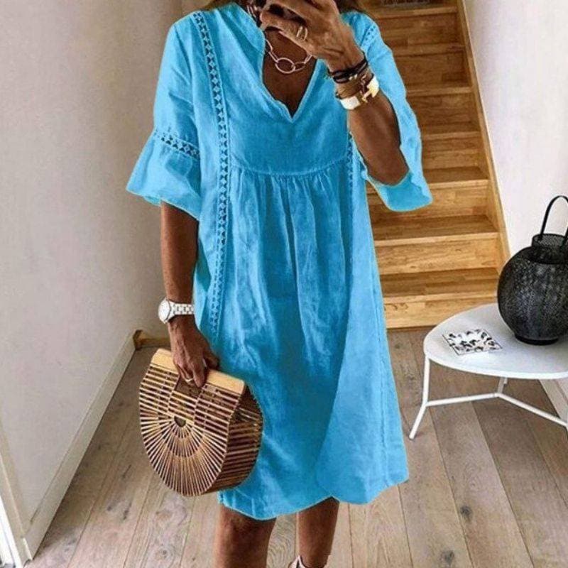 Frau in blauem Sommerkleid mit V-Ausschnitt und Rüschenärmeln, hält eine Bambustasche. Steht in modernem Innenraum mit Holztreppe.