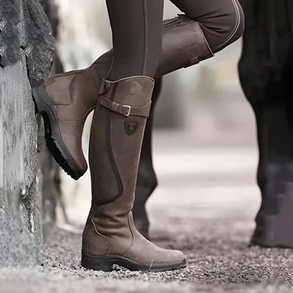 Braune Reitstiefel aus Leder, getragen von einer Person in brauner Reithose, lehnt an einer Steinmauer. Outdoor-Reitbekleidung, robustes Design.