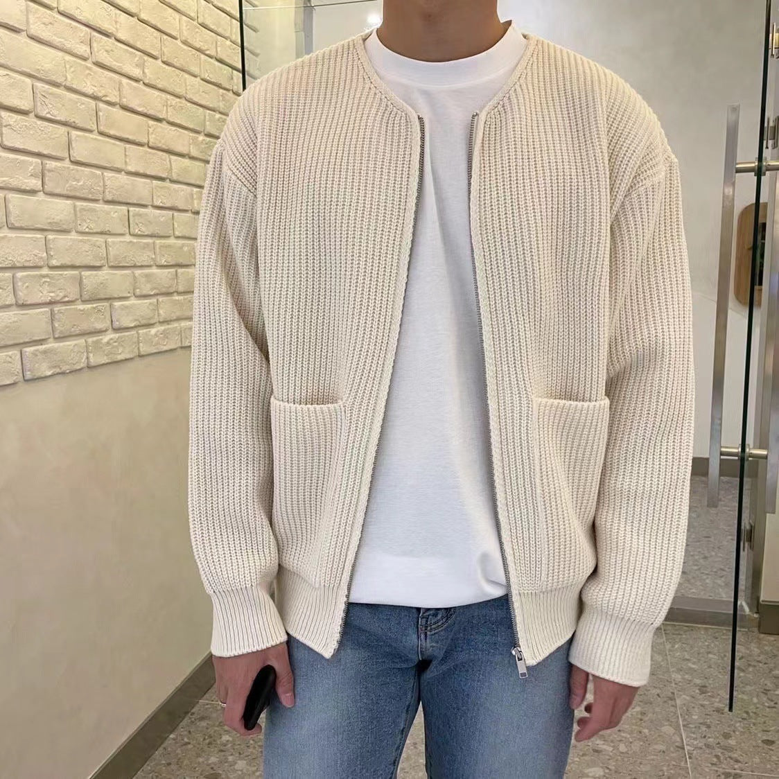 Mann in cremefarbener Strickjacke und weißem T-Shirt, blaue Jeans, steht vor Ziegelwand. Modetrend, lässiger Herrenstil, Strickmode.