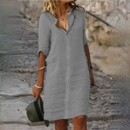 Frau in grauem Leinenkleid auf Kopfsteinpflaster, hält Hut in der Hand. Sommermode, lässiger Stil, elegantes Freizeitoutfit.
