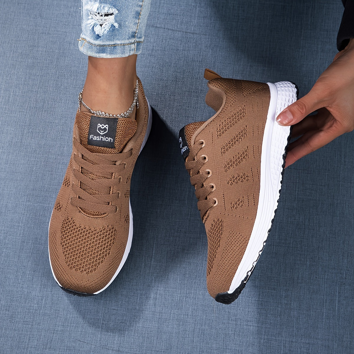 Braune Sportschuhe mit weißer Sohle, gestricktem Obermaterial und Fashion-Logo, getragen von Person in zerrissener Jeans auf blauem Hintergrund.