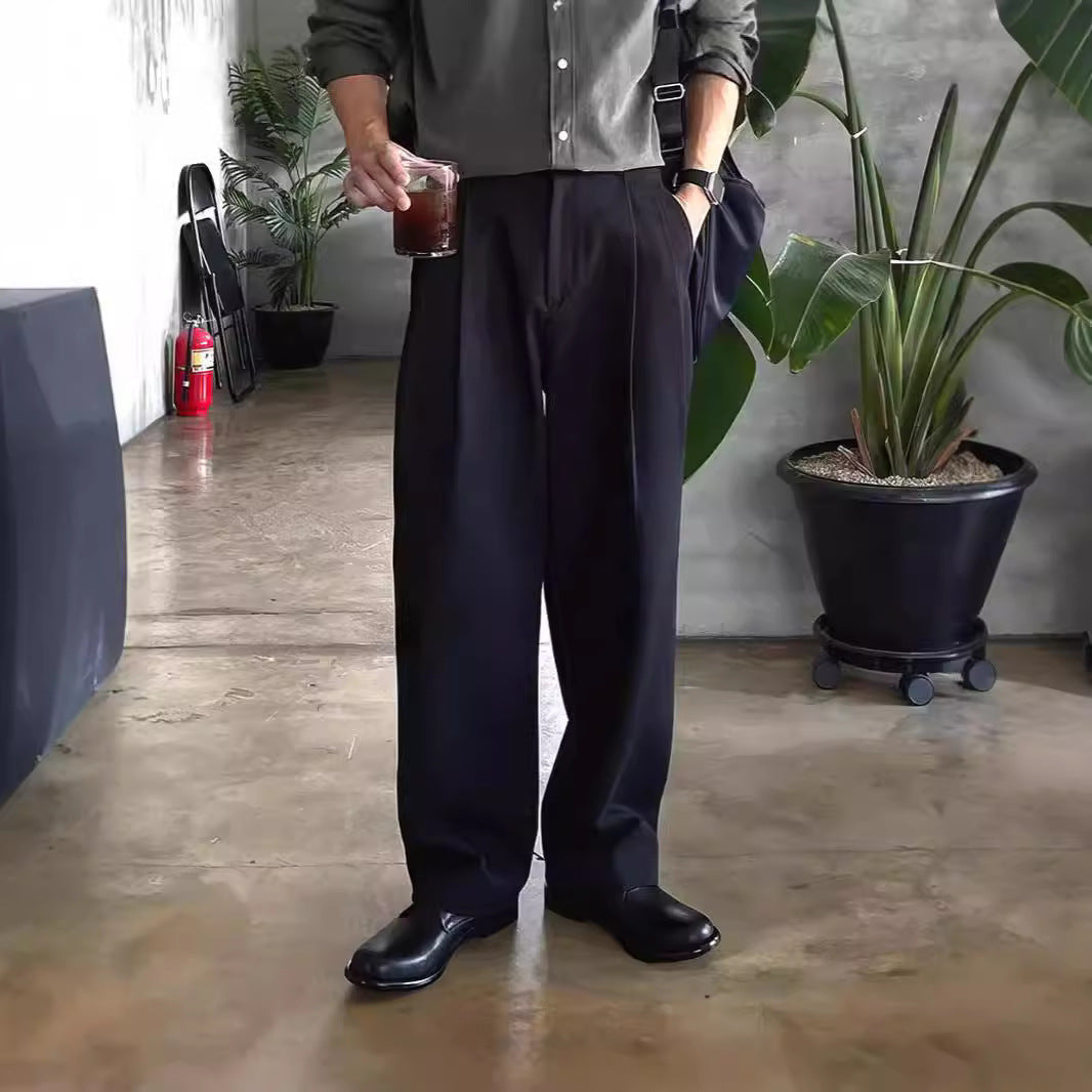 Mann in schwarzer Hose und Hemd hält Getränk, steht neben Pflanze in modernem Raum. Elegante Herrenmode, lässiger Stil, Indoor-Pflanzen.
