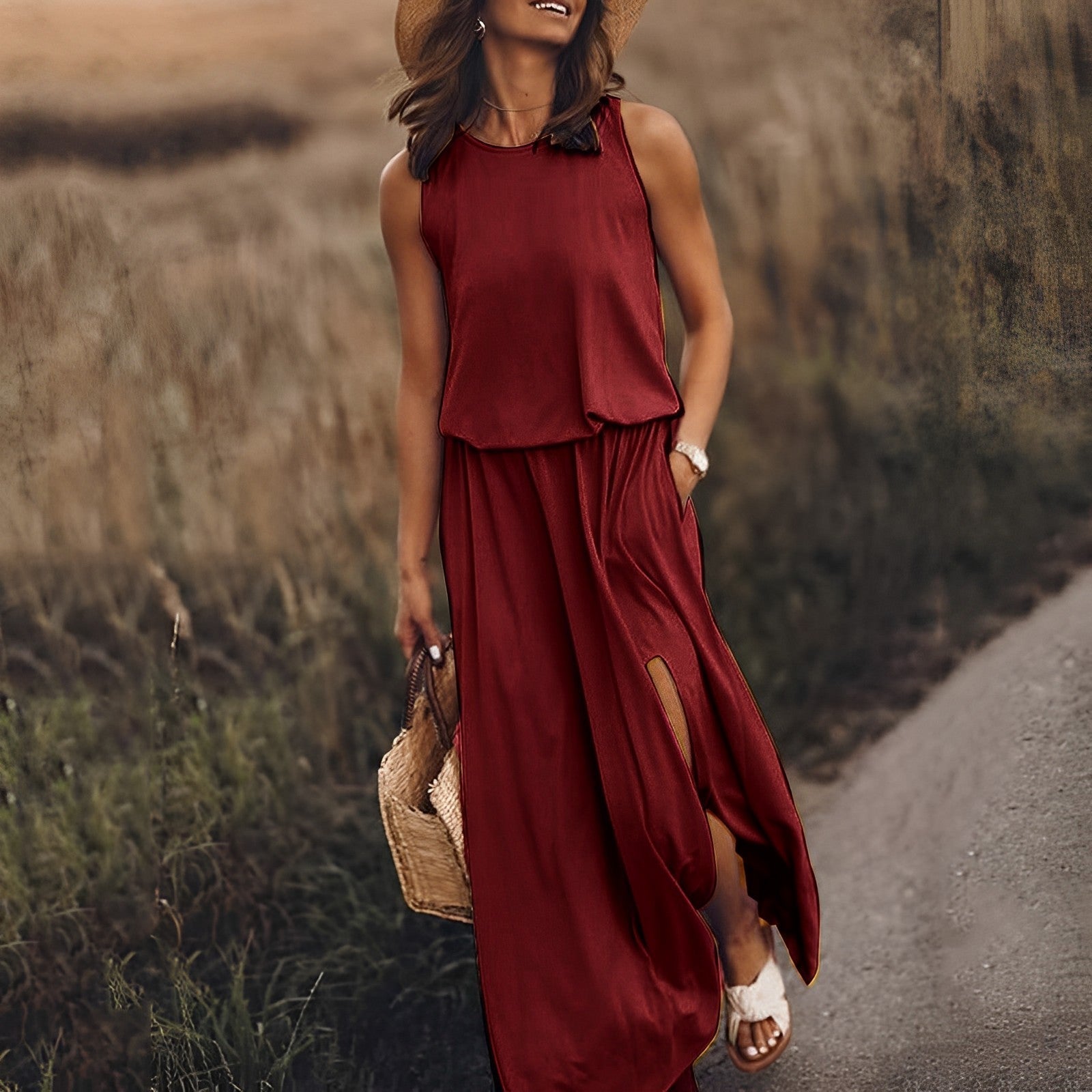 Frau in rotem Maxikleid mit Schlitz, Strohhut und Korbtasche spaziert auf ländlichem Weg. Sommermode, eleganter Freizeitlook, Naturkulisse.