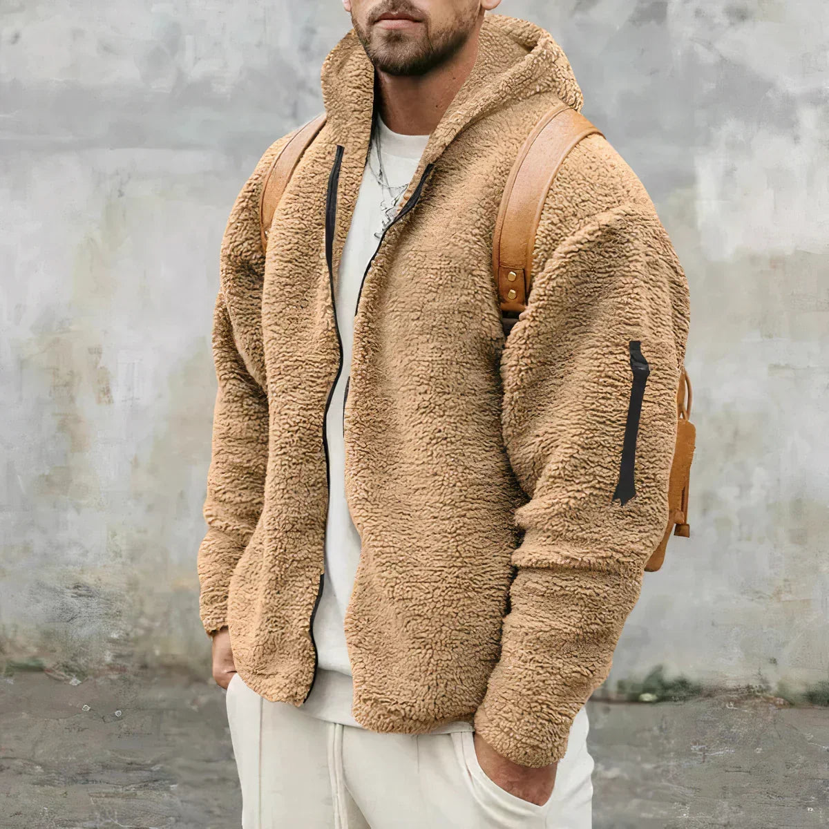 Mann in beiger Teddyfelljacke mit Kapuze und braunem Rucksack vor grauer Wand. Modetrend, Winterjacke, Herrenmode, Outdoor-Bekleidung.