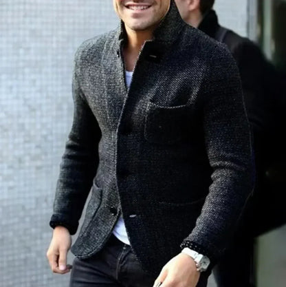 Ein Mann in einem eleganten, grauen Wollblazer mit weißem Hemd und Armbanduhr, lächelnd, vor einer modernen, strukturierten Wand.