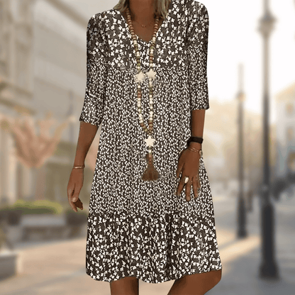 Frau in geblümtem Sommerkleid mit langen Ketten, spaziert auf sonniger Stadtstraße. Modetrend, Damenmode, Freizeitkleid, urbaner Stil.