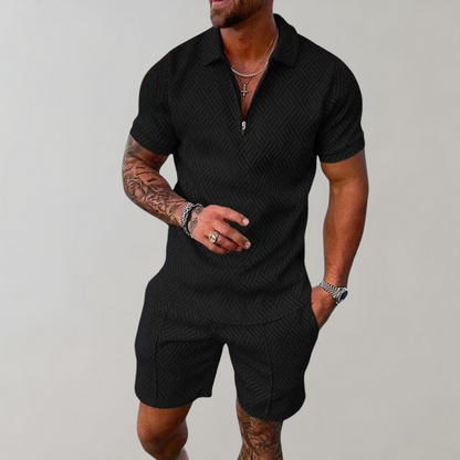 Schwarzer Herren-Shorts-Overall mit geometrischem Muster, getragen von muskulösem Mann mit Tattoos, silbernen Ketten und Armbändern vor grauem Hintergrund.