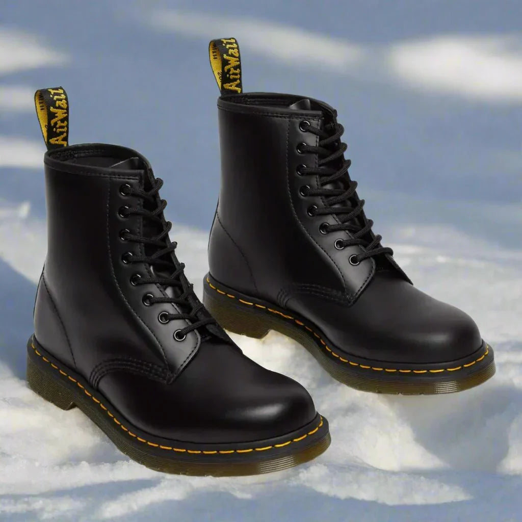 Schwarze Dr. Martens Stiefel im Schnee, ikonisches Design mit gelber Naht und AirWair-Schlaufe, robuste Winterstiefel, modisch und langlebig.