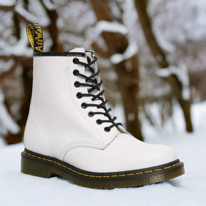Weiße Dr. Martens Stiefel im Schnee, klassische Schnürstiefel mit gelber Naht und markantem Logo, ideal für Wintermode und Outdoor-Aktivitäten.