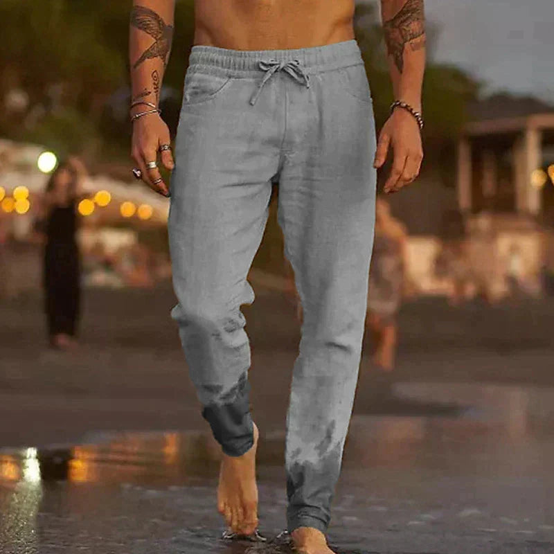 Ein Mann in grauer Jogginghose geht barfuß am Strand entlang. Abendstimmung, verschwommene Lichter im Hintergrund. Freizeitmode, Strandspaziergang.