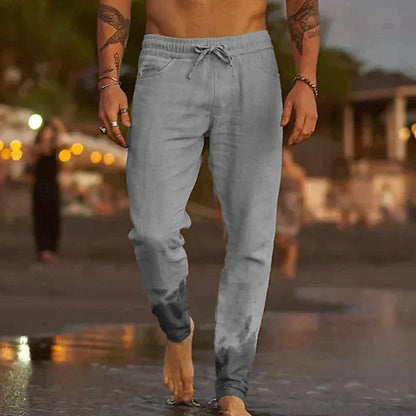 Ein Mann in grauer Jogginghose geht barfuß am Strand entlang. Abendstimmung, verschwommene Lichter im Hintergrund. Freizeitmode, Strandspaziergang.