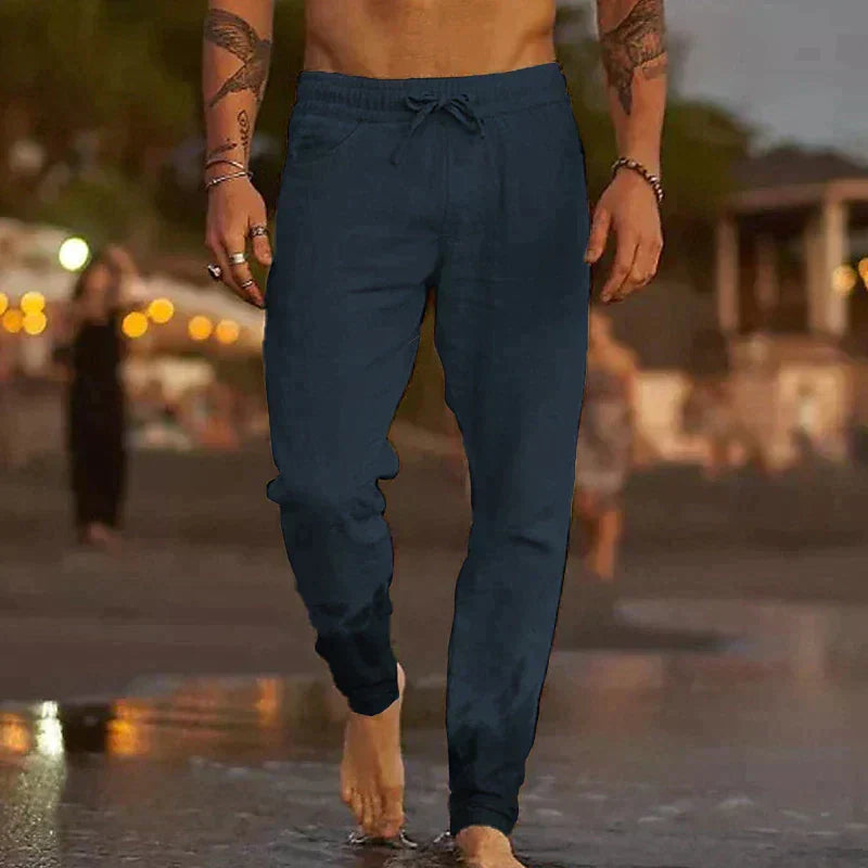 Mann in blauer Jogginghose am Strand bei Sonnenuntergang, barfuß im Wasser. Freizeitmode, Sommerabend, entspannte Atmosphäre.