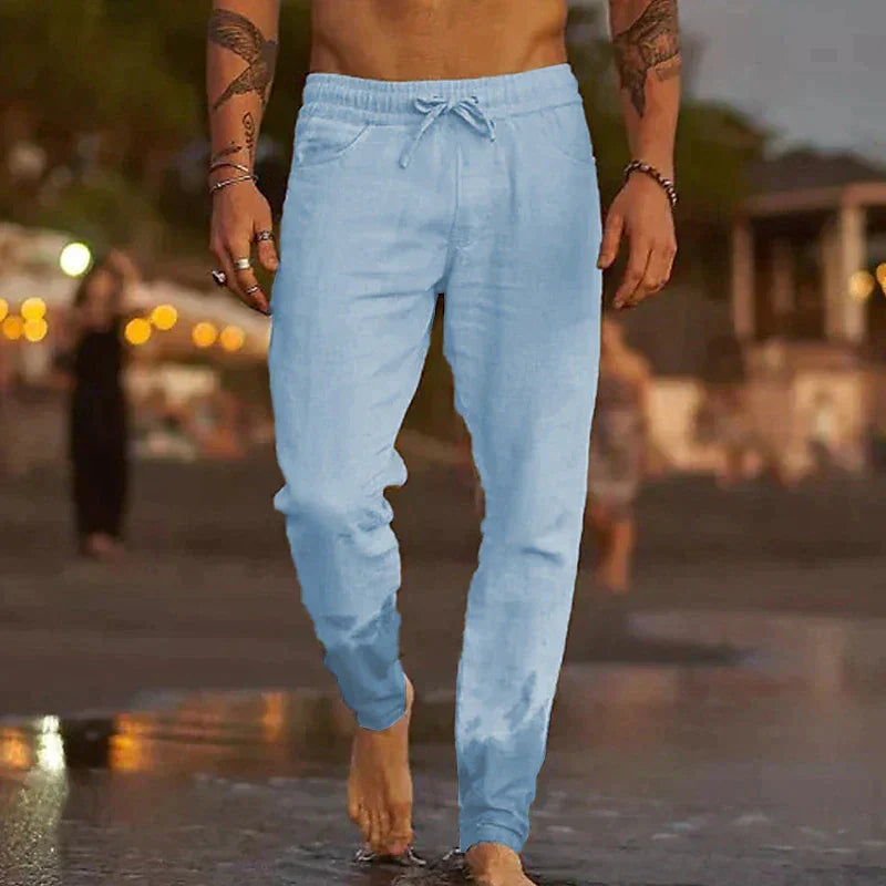 Mann in hellblauer Jogginghose am Strand bei Sonnenuntergang, barfuß im Wasser. Freizeitmode, Sommerkleidung, Strandspaziergang.
