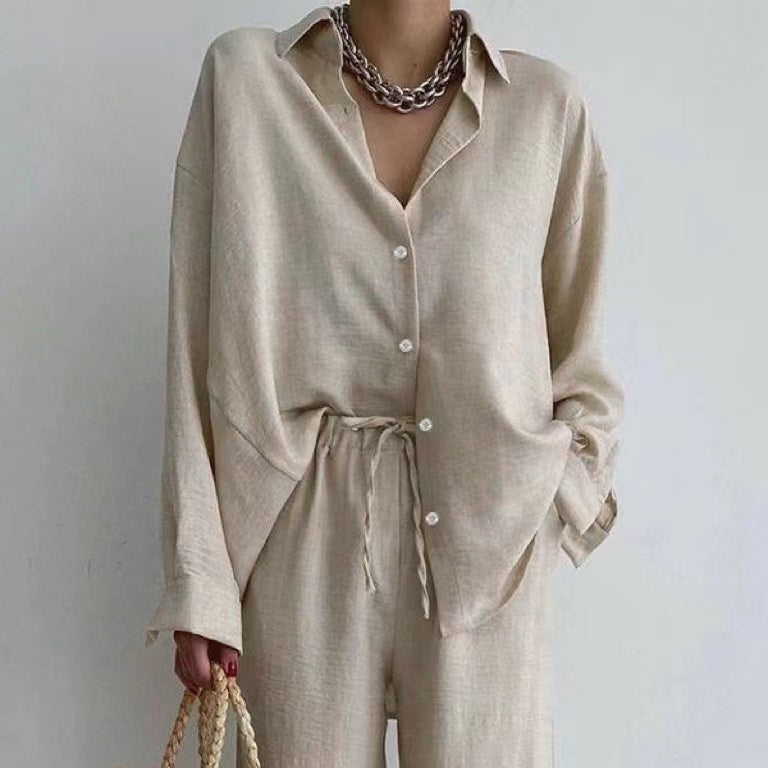 Elegante Frau in beigem Leinenhemd und Hose, trägt eine Kette und hält eine geflochtene Tasche. Minimalistischer, moderner Modestil.