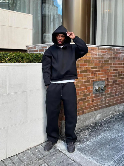 Mann in schwarzem Hoodie und Jogginghose steht vor Backsteinwand. Streetwear, urbaner Stil, lässige Mode, Herrenbekleidung, Stadtumgebung.