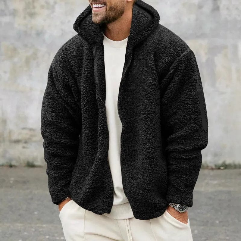 Mann in schwarzer Teddyfleece-Jacke und weißem Outfit, lächelnd, vor grauer Wand. Modetrend, Herrenbekleidung, Winterjacke, lässiger Stil.