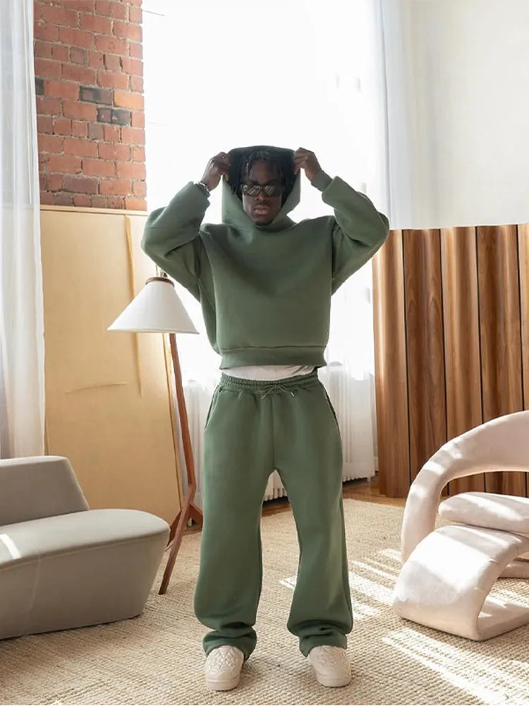 Person in grünem Hoodie und Jogginghose steht in modernem Wohnzimmer mit Stehlampe und Designerstuhl. Lässige Mode, gemütliche Einrichtung.