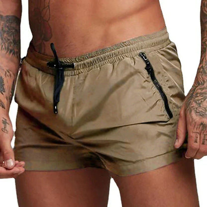 Mann mit tätowierten Armen trägt beige Badehose mit schwarzen Reißverschlusstaschen. Sommermode, Herrenbadebekleidung, Strandoutfit.