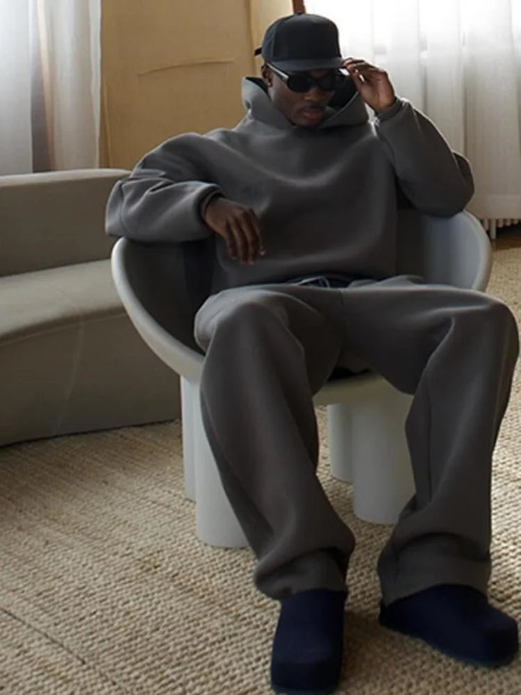 Ein Mann in grauem Oversize-Hoodie und Jogginghose sitzt auf einem modernen Stuhl, trägt eine schwarze Kappe und Sonnenbrille.