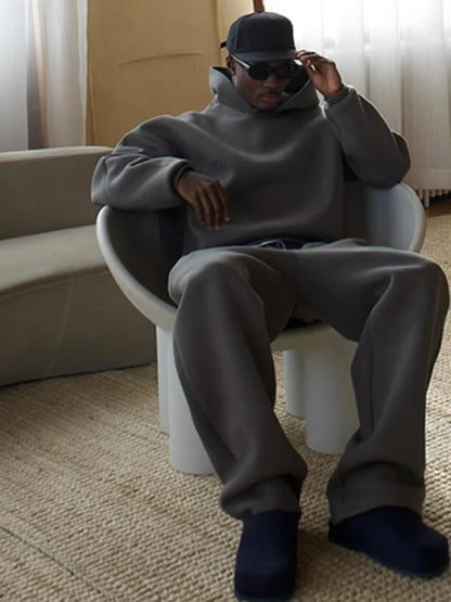 Ein Mann in grauem Oversize-Hoodie und Jogginghose sitzt auf einem modernen Stuhl, trägt eine schwarze Kappe und Sonnenbrille.