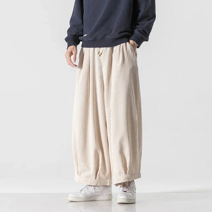 Person trägt weite, beige Haremshose und weiße Sneaker vor weißem Hintergrund. Modetrend, lässiger Stil, Herrenbekleidung, Streetwear.