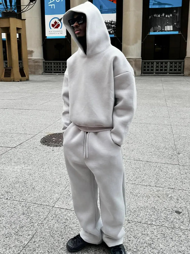 Person in grauem Hoodie und Jogginghose, trägt Sonnenbrille, steht auf städtischem Platz. Streetwear, lässiger Stil, urbaner Look.