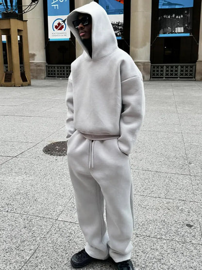 Person in grauem Hoodie und Jogginghose, trägt Sonnenbrille, steht auf städtischem Platz. Streetwear, lässiger Stil, urbaner Look.