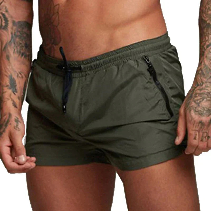 Mann in olivgrüner Badehose mit Taschen und Kordelzug, tätowierte Arme, sportlicher Körperbau. Ideal für Strandmode und Sommeraktivitäten.