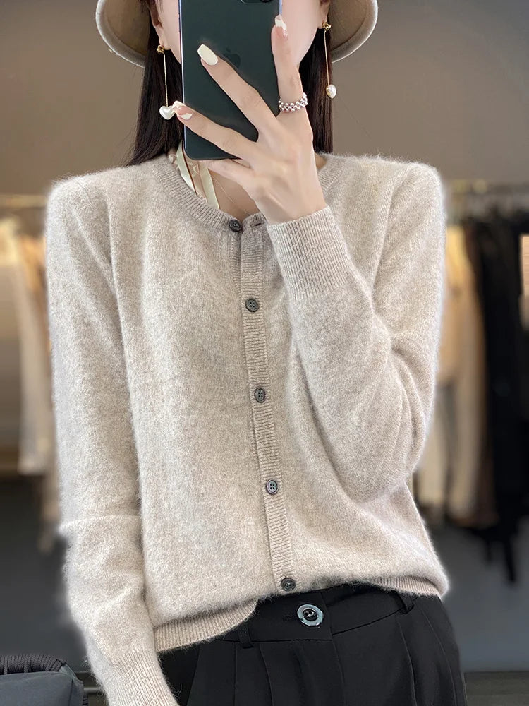 Dame in beigem Strickcardigan und schwarzer Hose, hält Smartphone, trägt Hut und Ohrringe. Modetrend, Herbstmode, eleganter Look.