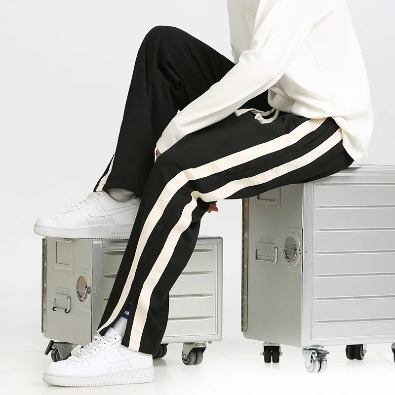 Weiße Sneakers und schwarze Jogginghose mit weißen Streifen, getragen von einer Person, die auf silbernen Metallboxen sitzt. Sportlich, modern, lässig.