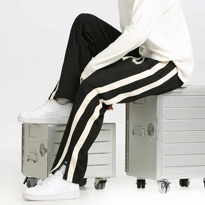 Weiße Sneakers und schwarze Jogginghose mit weißen Streifen, getragen von einer Person, die auf silbernen Metallboxen sitzt. Sportlich, modern, lässig.