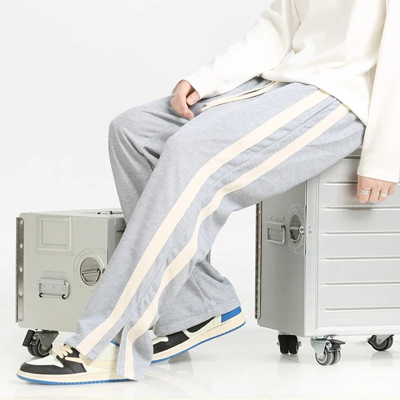 Graue Jogginghose mit weißen Streifen, getragen von einer Person, die auf silbernen Koffern sitzt. Bequeme Freizeitmode, ideal für Reisen.