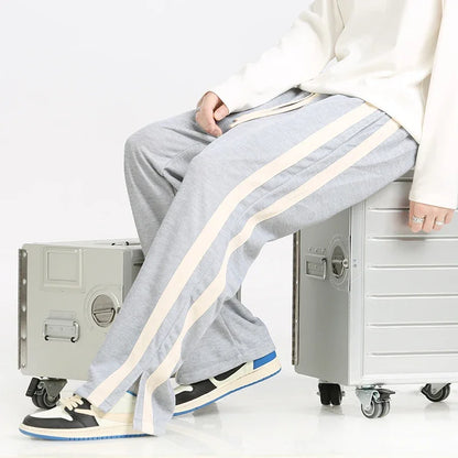 Graue Jogginghose mit weißen Streifen, getragen von einer Person, die auf silbernen Koffern sitzt. Bequeme Freizeitmode, ideal für Reisen.