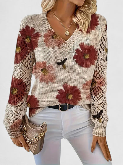 Frau trägt cremefarbenen Pullover mit roten Blumenmuster, V-Ausschnitt und gehäkelten Ärmeln, kombiniert mit weißer Hose und beiger Handtasche.