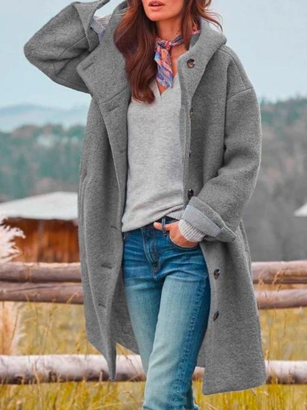 Frau in grauem Wollmantel, Jeans und Schal steht lässig vor ländlicher Kulisse. Herbstmode, Outdoor, stilvoll gekleidet.