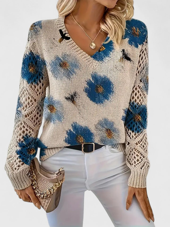 Frau trägt cremefarbenen Strickpullover mit blauen Blumen, V-Ausschnitt und Häkelärmeln, kombiniert mit weißer Hose und beiger Handtasche.